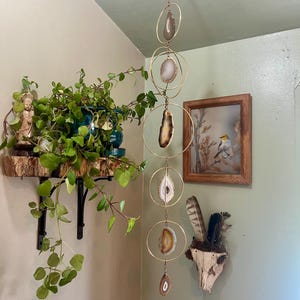 May include: A vertical wall hanging mobile featuring five circular gold-toned metal rings suspended by a thin chain. Each ring contains a polished, translucent agate slice in shades of brown, white, and gray. A small, teardrop-shaped agate slice hangs at the bottom.