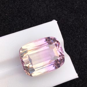 以下が含まれることがあります： パステルカラーの大きな長方形の宝石。ピンク、ピーチ、白の色合いが混ざり合い、光を反射するファセットカットが施されています。石は暗い背景に対して白い表面に置かれています。