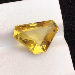 Könnte beinhalten: Ein facettierter, dreieckiger Edelstein in leuchtendem Gelb. Der Stein liegt auf einer weißen Oberfläche, mit einem dunklen Hintergrund. Der Schliff des Steins zeigt mehrere Facetten, die seine Brillanz verstärken. Dieser Edelstein ist für Schmuck geeignet.