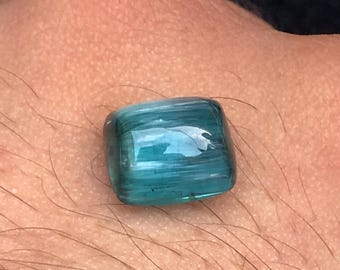 May include: A rectangular, teal-colored gemstone with a polished surface. The stone has a translucent quality, allowing light to pass through, and is held in a hand. The gemstone's color is a vibrant shade of blue-green.