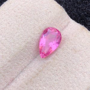 Puede incluir: Una gema rosa brillante en forma de pera y facetada. La gema está colocada sobre una tela rosa claro texturizada. Las facetas de la gema reflejan la luz, creando un efecto brillante. La presentación general sugiere un componente de joyería.