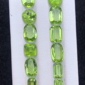 Könnte beinhalten: Eine Sammlung von Peridot-Edelsteinen in verschiedenen Formen, darunter oval, rund und Smaragdschliff. Die Edelsteine sind ein leuchtendes, durchscheinendes Grün und werden auf einer weißen Oberfläche präsentiert. Die Anordnung zeigt die verschiedenen Facetten und die Klarheit der Steine.