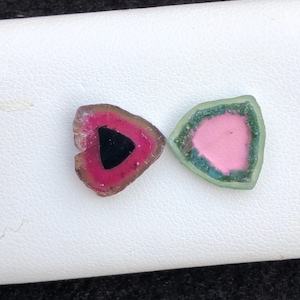 Könnte beinhalten: Zwei Wassermelonen-Turmalin-Edelsteine, die jeweils so geschnitten sind, dass sie ein einzigartiges Farbmuster zeigen. Einer hat einen schwarzen Mittelteil, der von Rosa und Grün umgeben ist, der andere einen rosa Mittelteil mit grünen und blauen Rändern. Die Steine ​​werden auf einer weißen Oberfläche ausgestellt.