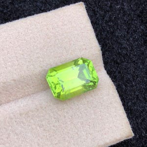 Könnte beinhalten: Ein rechteckiger, facettierter Peridot-Edelstein in leuchtendem, hellgrün. Der Stein liegt auf einer hellbeigen Oberfläche, die sich von dem dunklen, strukturierten Hintergrund abhebt. Der Schliff des Steins lässt das Licht reflektieren.