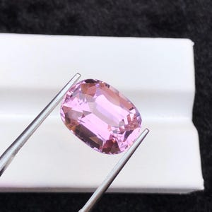 Könnte beinhalten: Ein hellrosa, kissenförmiger Edelstein, der von einer Pinzette gehalten wird. Der Edelstein hat eine rechteckige Form mit abgerundeten Ecken und Facetten, die das Licht einfangen. Der Hintergrund ist eine dunkle, strukturierte Oberfläche.