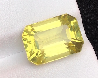 Puede incluir: Una gema facetada, de corte rectangular, con un tono amarillo vibrante. La gema se encuentra sobre una superficie blanca, mostrando su claridad y brillo. El corte presenta múltiples facetas que reflejan la luz. Ideal para joyería.