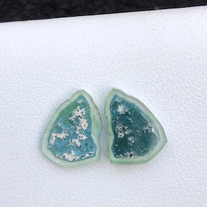 Könnte beinhalten: Zwei unregelmäßig geformte, durchscheinende Glasstücke in Hellblau- und Grüntönen. Das Glas hat ein strukturiertes Aussehen mit weißen Einschlüssen, und die Ränder sind heller grün.