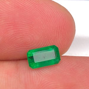 Könnte beinhalten: Ein leuchtend grüner, rechteckig geschliffener Smaragd. Der Edelstein hat eine polierte Oberfläche und wird zwischen zwei Fingern gehalten. Der Smaragd ist von lebendiger Farbe. Dieser Edelstein eignet sich für die Schmuckherstellung.