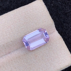 Könnte beinhalten: Ein rechteckiger, hellvioletter Edelstein mit Facettenschliff. Der Edelstein liegt auf einer hellbeigen Oberfläche mit dunklem Hintergrund. Die Facetten des Edelsteins reflektieren das Licht. Geeignet für die Schmuckherstellung.