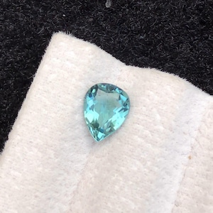 Könnte beinhalten: Ein birnenförmiger, hellblauer Edelstein liegt auf einem weißen Tuch vor einem dunklen Hintergrund. Der Edelstein hat eine facettierte Oberfläche, die das Licht reflektiert. Die Farbe des Edelsteins ist ein leuchtendes, klares Türkis.