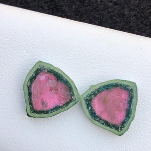 Puede incluir: Dos rodajas de piedras preciosas de turmalina sandía. Cada rodaja tiene un centro rosa vibrante, un borde exterior verde oscuro y un borde delgado de color verde oscuro. Las piedras preciosas se exhiben sobre una superficie blanca.