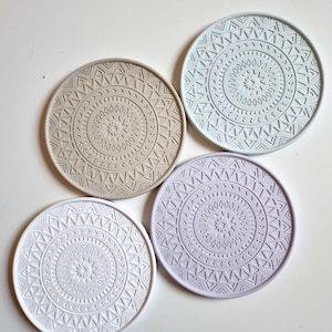 Puede incluir: Cuatro posavasos redondos con un patrón geométrico. Los posavasos son en tonos de gris, azul claro y morado.