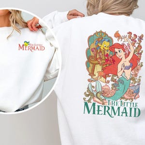 May include: White sweatshirt featuring a colorful graphic of Ariel and Flounder from "The Little Mermaid" on the back. The front left chest has a small embroidered logo. The text "The Little Mermaid" is also displayed.