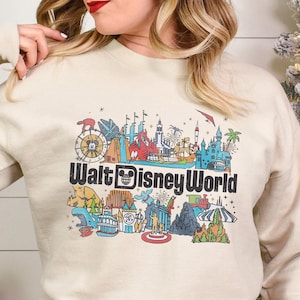 Puede incluir: Sudadera de color arena con un gr&aacute;fico colorido de las atracciones de Walt Disney World. El dise&ntilde;o incluye una noria, un castillo y otros monumentos ic&oacute;nicos. El texto "Walt Disney World" se muestra en negro.