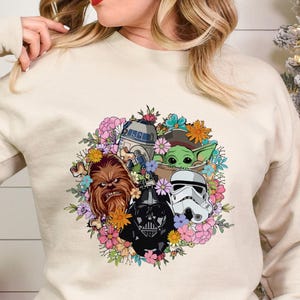 Puede incluir: Sudadera color arena con un dise&ntilde;o de corona floral con personajes de Star Wars, incluyendo a Chewbacca, Darth Vader, un Stormtrooper, R2-D2 y Grogu. La sudadera tiene mangas largas y cuello redondo.