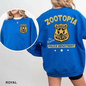 Pode incluir: Camisola azul royal com um design do Departamento de Pol&iacute;cia de Zootopia. A frente tem um gr&aacute;fico de distintivo, enquanto a parte de tr&aacute;s exibe o logotipo e o distintivo de Zootopia, com o texto "Now Recruiting". Um item de vestu&aacute;rio divertido e tem&aacute;tico.