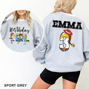 May include: A light gray sweatshirt with the text "Birthday Girl" and cartoon characters on the front. The back features the name "EMMA" and a number "3" with a cowgirl hat and braid design. The sweatshirt is labeled "SPORT GREY".