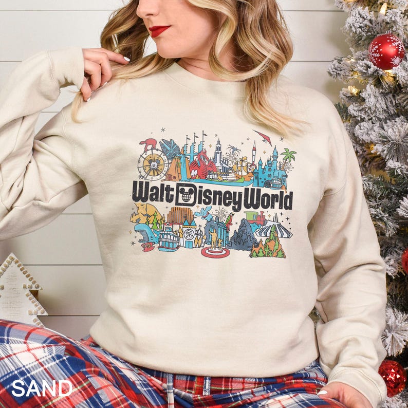 Puede incluir: Sudadera de color arena con un gr&aacute;fico colorido de las atracciones de Walt Disney World. El dise&ntilde;o incluye una noria, un castillo y otros monumentos ic&oacute;nicos. El texto "Walt Disney World" se muestra en negro.