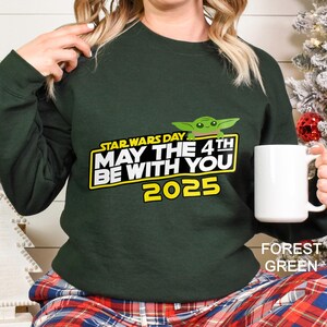 May include: Forest green sweatshirt with the text "STAR WARS DAY MAY THE 4TH BE WITH YOU 2025" in yellow and white, and a green cartoon character. A white mug is held in the right hand.