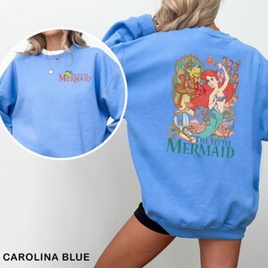 Op de afbeelding: Carolina blauwe sweatshirt met de tekst "The Little Mermaid" in groen en rood. De voorkant heeft een klein logo en de achterkant toont een kleurrijke afbeelding van Ariel, Botje en Sebastiaan. Een casual, comfortabele top.