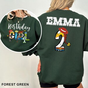 May include: Forest green sweatshirt with white trim featuring the text "Birthday Girl" and Toy Story characters. The back displays "EMMA" and a number 2 with a cowgirl hat and braid design. A fun, festive birthday garment.