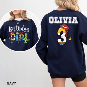 May include: Navy blue sweatshirt with "Birthday Girl" and character graphics on the front. The back features the name "Olivia" and a number "3" with a cowgirl design. The sweatshirt is a comfortable, casual style.
