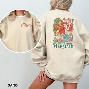 Op de afbeelding: Zandkleurige sweatshirt met een kleurrijke afbeelding van Ariel, Botje en Sebastiaan uit "De Kleine Zeemeermin" op de achterkant. De linkerborst heeft de woorden "The Little Mermaid". Het woord "SAND" staat onderaan.