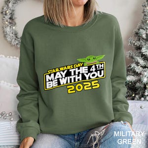 May include: An olive green sweatshirt featuring the text "STAR WARS DAY MAY THE 4TH BE WITH YOU 2025" in yellow and white, with a green cartoon character. The sweatshirt is a crew neck style.