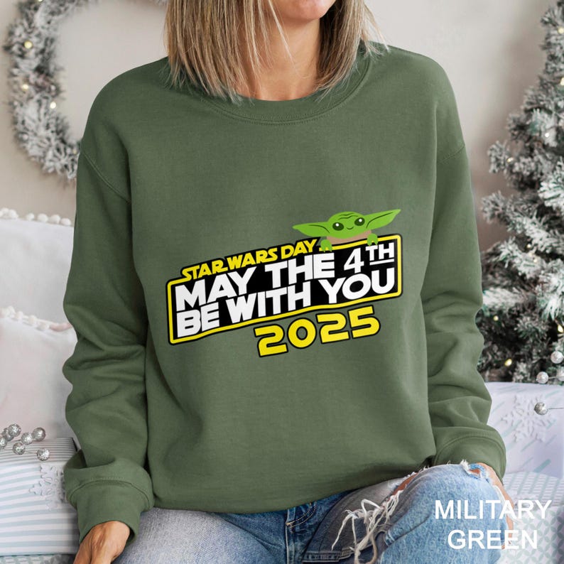 May include: An olive green sweatshirt featuring the text "STAR WARS DAY MAY THE 4TH BE WITH YOU 2025" in yellow and white, with a green cartoon character. The sweatshirt is a crew neck style.