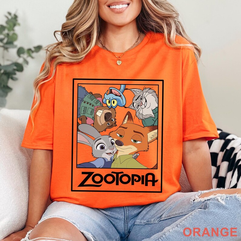 Peut inclure: T-shirt orange vif avec un motif du film d'animation Zootopie. Le design pr&eacute;sente des personnages du film, avec le mot "Zootopia" en noir en bas de l'image.