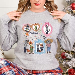 Pode incluir: Camisola cinzenta desportiva com personagens de Zootopia. O design inclui imagens de desenhos animados de uma raposa, um coelho e outras personagens em molduras e cora&ccedil;&otilde;es. A palavra "Zootopia" est&aacute; escrita numa fonte estilizada, com a frase "welcome to zootopia" por baixo.