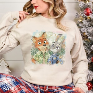 May include: A sand-coloured sweatshirt with a colourful graphic of Nick Wilde and Judy Hopps from Zootopia. Nick is wearing a green Hawaiian shirt, and Judy is in a police uniform. The word "SAND" is printed at the bottom.