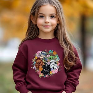 Puede incluir: Una sudadera granate con un dise&ntilde;o floral con personajes de Star Wars, incluyendo Darth Vader, Chewbacca y un Stormtrooper. El gr&aacute;fico est&aacute; rodeado de flores coloridas. La sudadera la lleva una persona joven con pelo largo casta&ntilde;o.