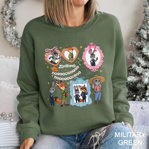 Pode incluir: Camisola verde militar com gola redonda e ilustra&ccedil;&otilde;es de personagens de Zootopia. O design inclui Judy Hopps, Nick Wilde e outros personagens em molduras decorativas. A palavra "Zootopia" est&aacute; escrita em uma fonte l&uacute;dica, rodeada por elementos decorativos.