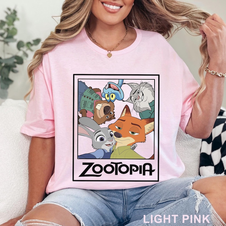 Peut inclure: T-shirt rose clair avec un graphisme encadr&eacute; du film d'animation "Zootopie". Le graphisme comprend des personnages comme Judy Hopps et Nick Wilde. Le mot "ZOOTOPIA" est imprim&eacute; en noir en dessous. Le t-shirt est rose clair.