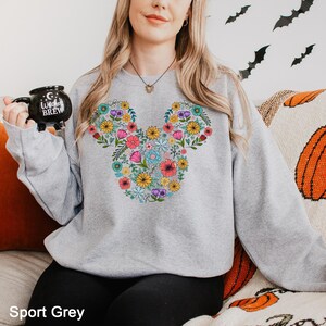 Puede incluir: Una sudadera gris claro con un dise&ntilde;o floral de Mickey Mouse. El dise&ntilde;o est&aacute; formado por varias flores coloridas. El texto "Sport Grey" est&aacute; en la parte inferior de la imagen. La persona sostiene una taza negra que dice "Witches Brew".