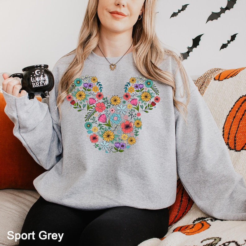 Puede incluir: Una sudadera gris claro con un dise&ntilde;o floral de Mickey Mouse. El dise&ntilde;o est&aacute; formado por varias flores coloridas. El texto "Sport Grey" est&aacute; en la parte inferior de la imagen. La persona sostiene una taza negra que dice "Witches Brew".