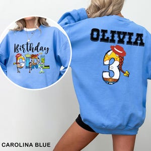 May include: Carolina blue sweatshirt featuring the text "Birthday Girl" with cartoon characters. The back of the sweatshirt displays the name "Olivia" above a number "3" with a cartoon design. The sweatshirt is a casual, comfortable style.