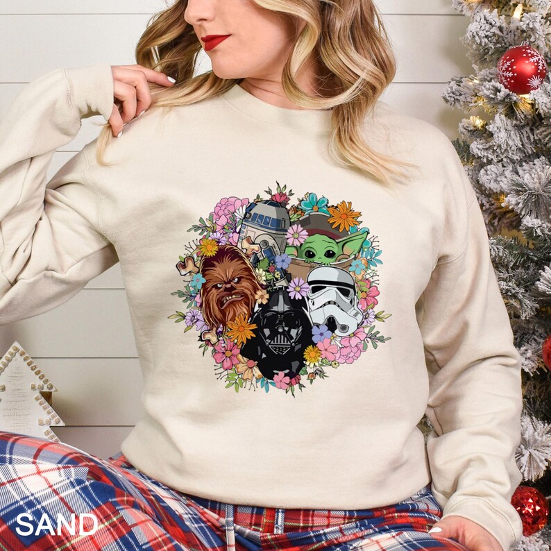 Puede incluir: Sudadera color arena con un dise&ntilde;o de corona floral con personajes de Star Wars, incluyendo a Chewbacca, Darth Vader, un Stormtrooper, R2-D2 y Grogu. La sudadera tiene mangas largas y cuello redondo.