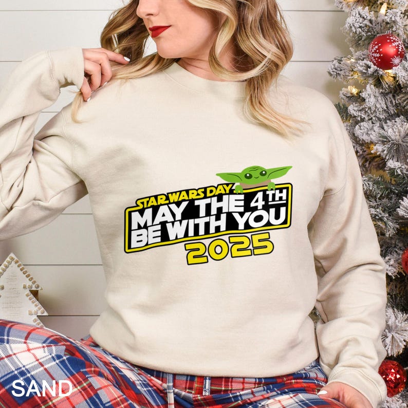 May include: Sand-colored sweatshirt featuring the text "STAR WARS DAY MAY THE 4TH BE WITH YOU 2025" in yellow and white, with a green cartoon character. The sweatshirt has long sleeves.