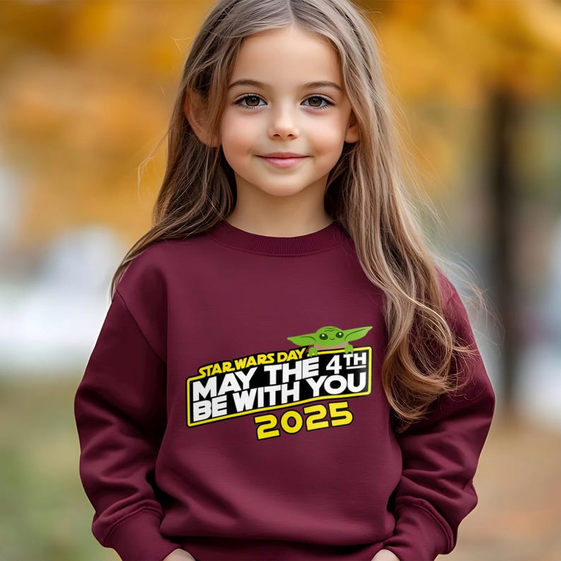 May include: A maroon sweatshirt featuring the text "STAR WARS DAY MAY THE 4TH BE WITH YOU 2025" with a green Baby Yoda graphic. The sweatshirt is worn by a young girl with long brown hair.