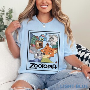 Peut inclure: T-shirt bleu clair avec un motif rectangulaire repr&eacute;sentant les personnages du film d'animation "Zootopie". Le motif comprend Judy Hopps, Nick Wilde et d'autres personnages. Le mot "Zootopie" est imprim&eacute; en noir. Le t-shirt est bleu clair.