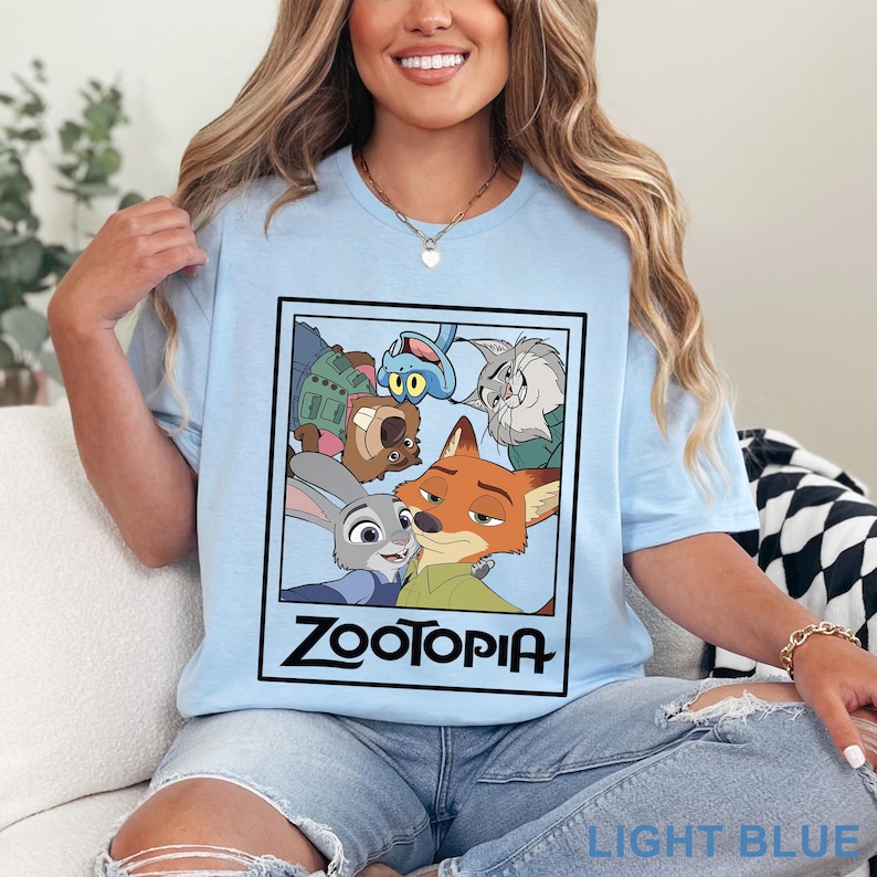 Peut inclure: T-shirt bleu clair avec un motif rectangulaire repr&eacute;sentant les personnages du film d'animation "Zootopie". Le motif comprend Judy Hopps, Nick Wilde et d'autres personnages. Le mot "Zootopie" est imprim&eacute; en noir. Le t-shirt est bleu clair.
