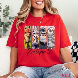 Peut inclure: T-shirt rouge avec des personnages de Zootopia 2. Le design comprend Nick Wilde, Judy Hopps, un gorille et Flash le paresseux, chacun dans un panneau vertical. Les mots "Zootopia 2" sont &eacute;crits en cursive en dessous.