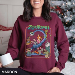 May include: Maroon sweatshirt featuring a colorful graphic from the movie Zootopia. The design includes various animal characters and the movie title in green. The sweatshirt is a solid maroon color.