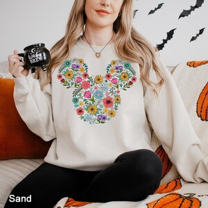 Puede incluir: Sudadera color arena con un dise&ntilde;o floral de Mickey Mouse en colores vibrantes. La sudadera tiene cuello redondo y mangas largas. La imagen tambi&eacute;n muestra una taza negra con el texto "Witches Brew".