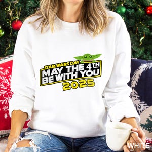 May include: White sweatshirt featuring the text "STAR WARS DAY MAY THE 4TH BE WITH YOU 2025" in yellow and black, with a green Grogu graphic. The sweatshirt is a long-sleeved top.