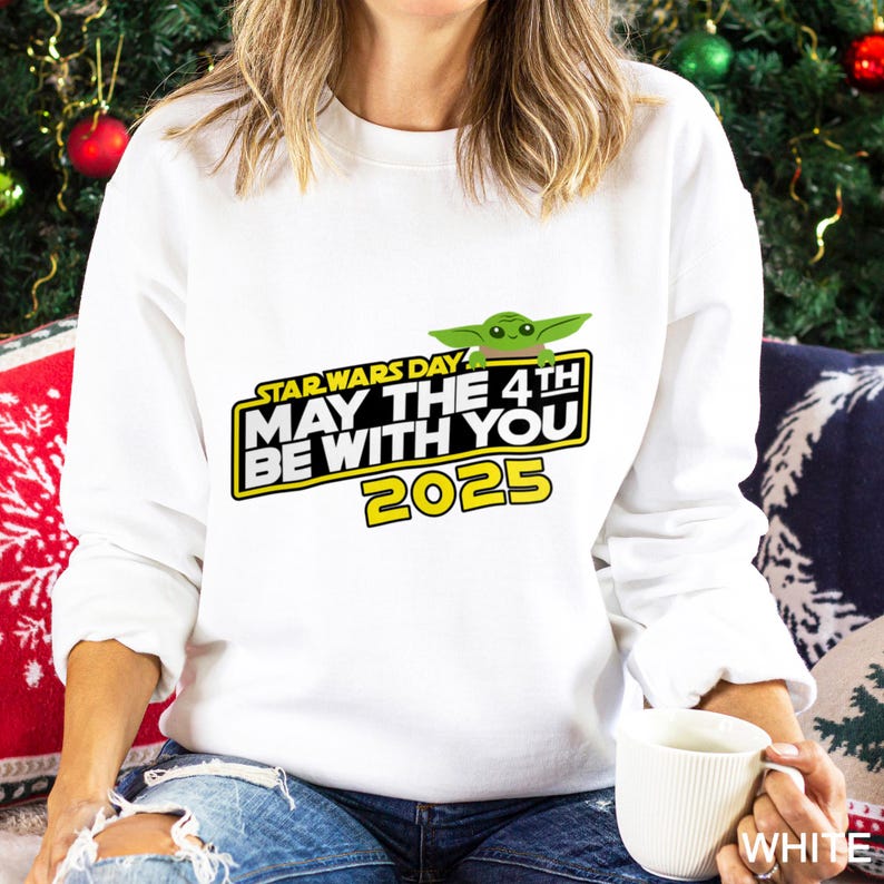 May include: White sweatshirt featuring the text "STAR WARS DAY MAY THE 4TH BE WITH YOU 2025" in yellow and black, with a green Grogu graphic. The sweatshirt is a long-sleeved top.