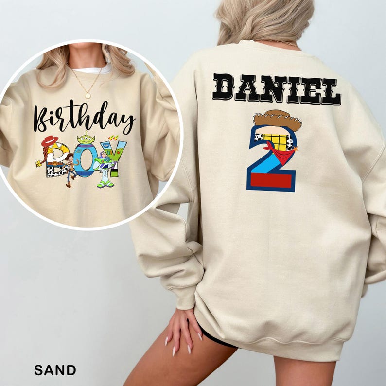 May include: Sand-colored sweatshirt with the text "Birthday Boy" and Toy Story characters on the front. The back features the name "Daniel" and a number "2" with a cowboy hat and bandana design. The sweatshirt is a casual, long-sleeved garment.
