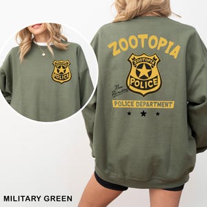 Pode incluir: Camisola verde militar com um distintivo do Departamento de Pol&iacute;cia de Zootopia na frente e atr&aacute;s. O design nas costas inclui a palavra "Zootopia" acima do distintivo e "Police Department" abaixo. A camisola tem mangas compridas.
